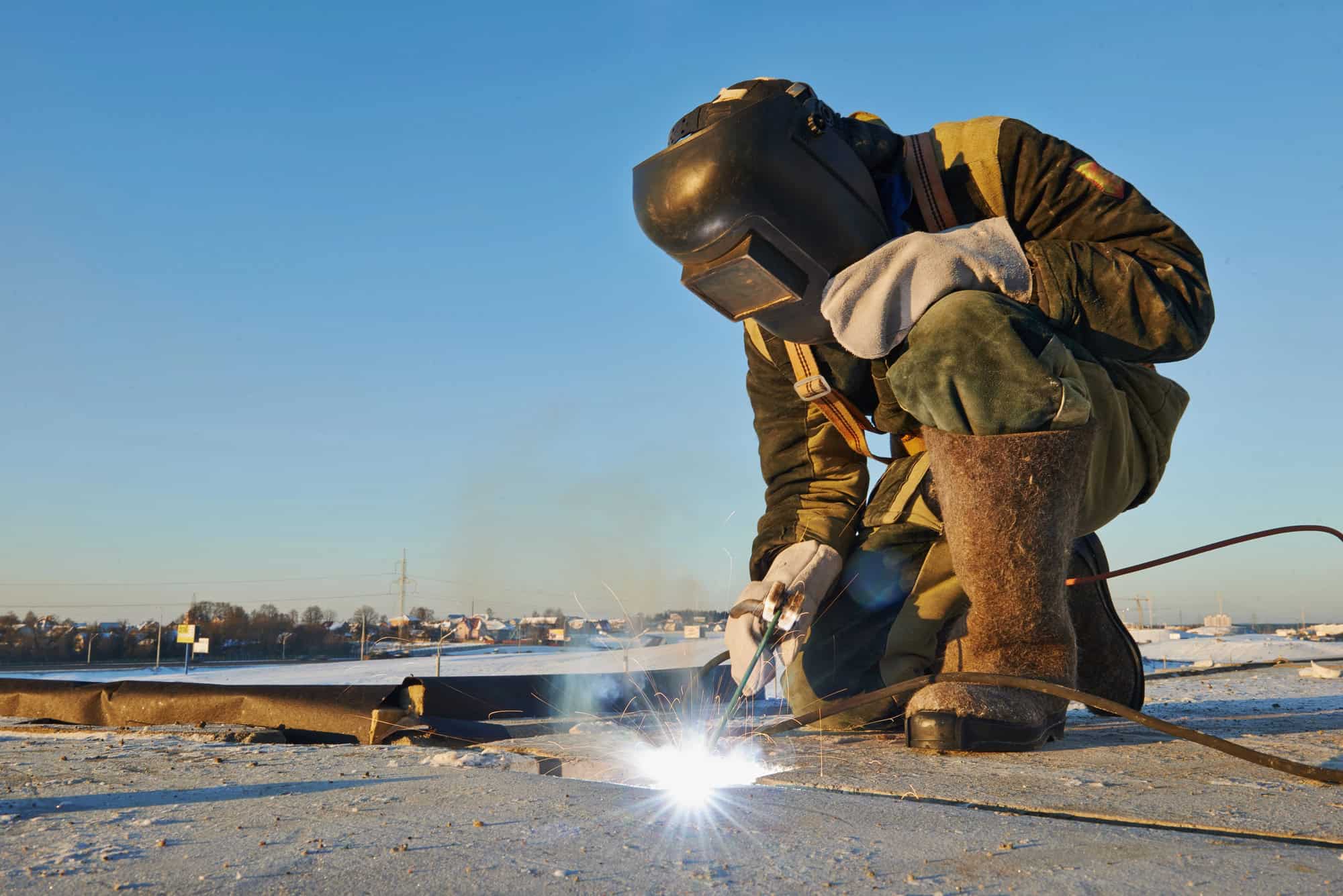 A person in protective gear welds metal outdoors on a sunny day.