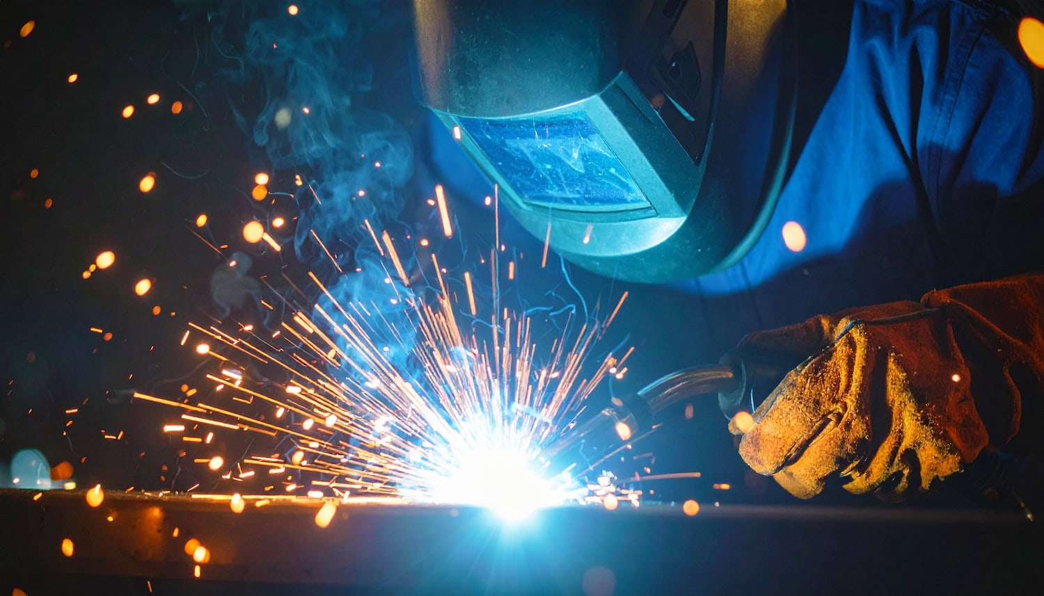 Person welding metal with protective gear, creating bright sparks and blue light.
