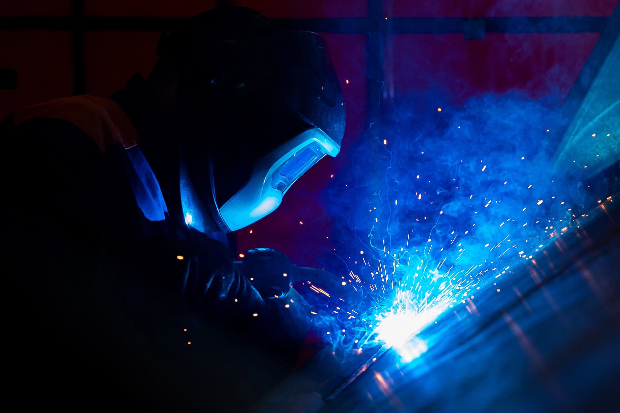 A person in protective gear welds metal, with blue sparks and smoke visible.