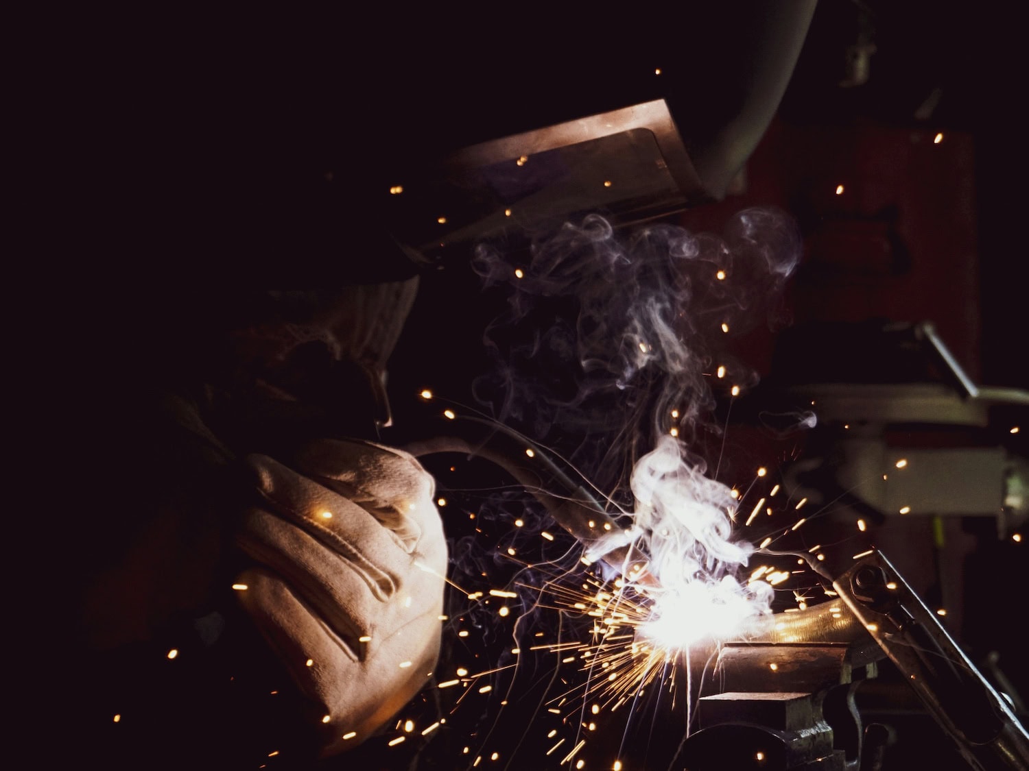A welder wearing gloves works with flying sparks and smoke around the welding area.