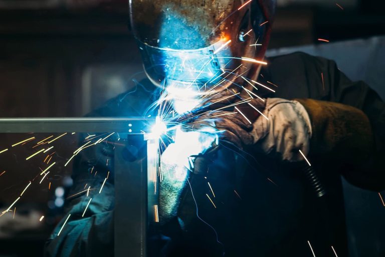 Person wearing a welding helmet welds metal with bright sparks and blue light.