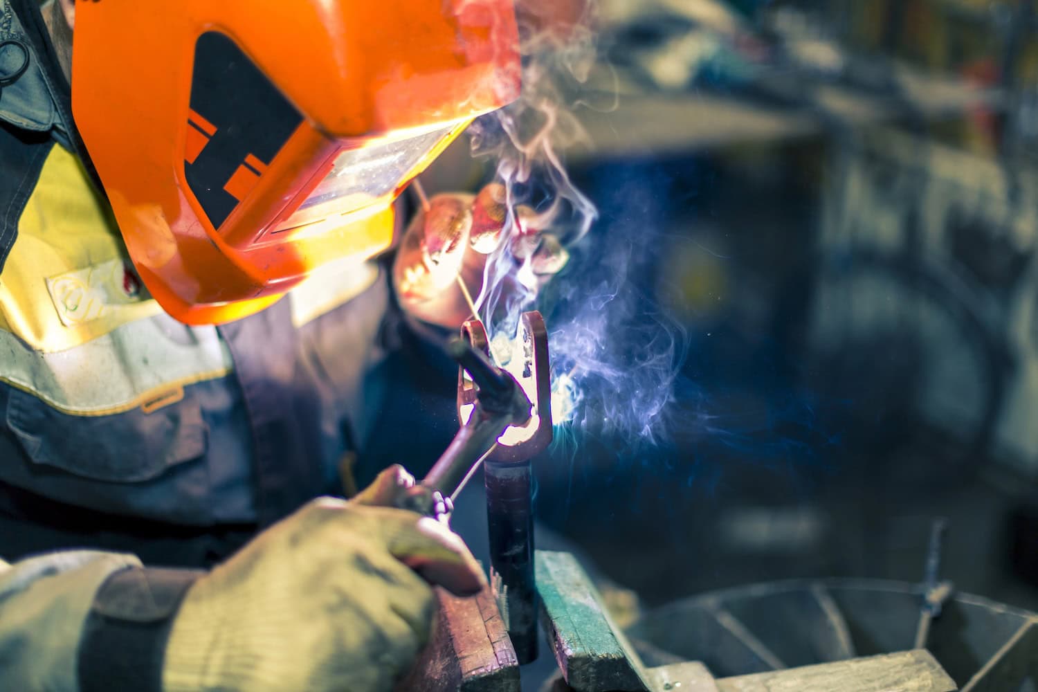 Person wearing protective gear welds metal with bright sparks and smoke visible.