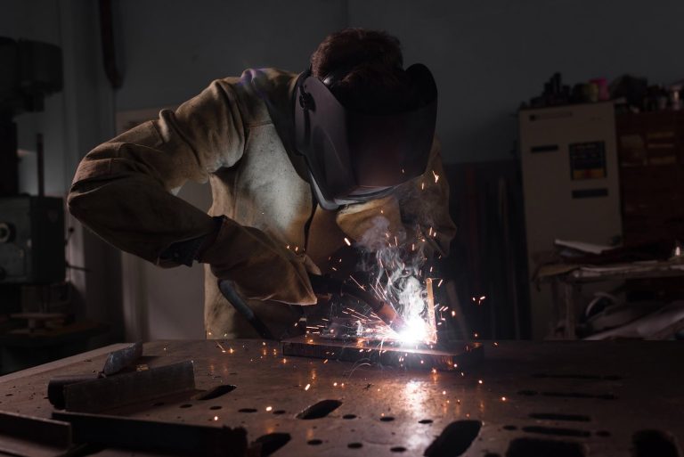 Person wearing protective gear welding metal, with sparks and smoke visible.